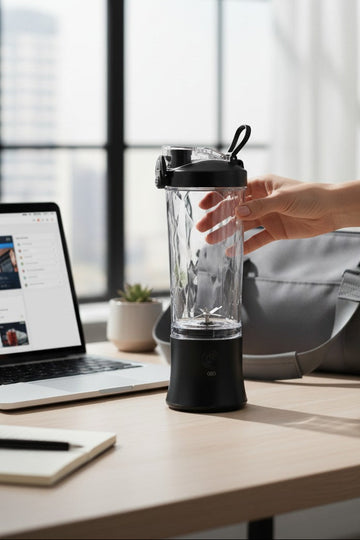 Hand holding a blender on a desk with a laptop and bag in the background