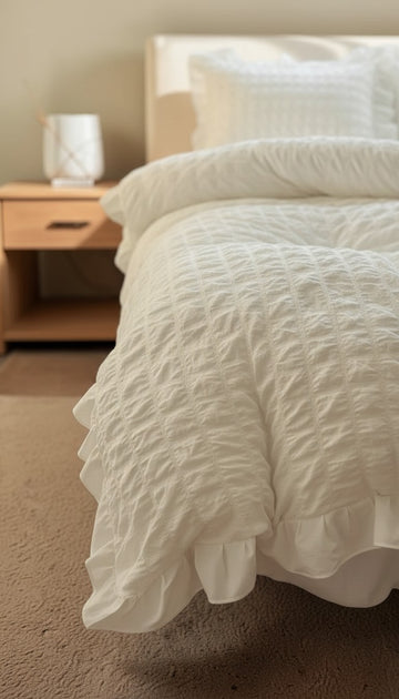 Close-up of a textured white comforter on a bed with a wooden nightstand in the background.